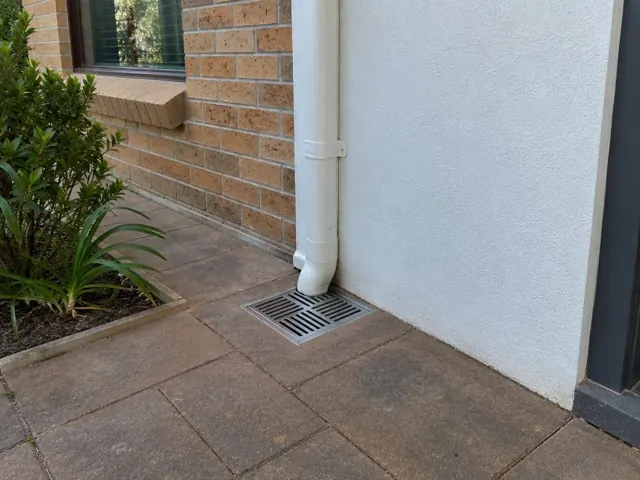 Outdoor drain gully with grate beside a brick and rendered house wall, paved path, and downpipe in Kellyville