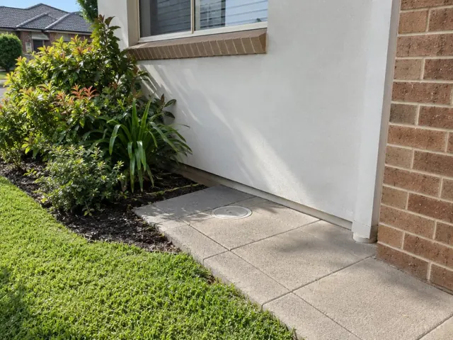 Flush circular access cover in paved side path beside a rendered house wall and garden in Beaumont Hills